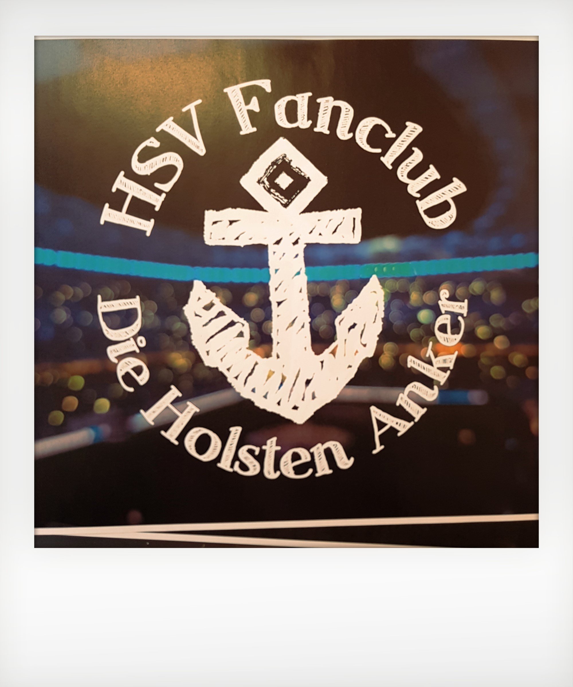 Foto: vorläufiges Logo HSV Fanclub. Mittig Anker, darum herum Schriftzug: HSV Fanclub Die Holsten Anker. Im Hintergrund Stadion