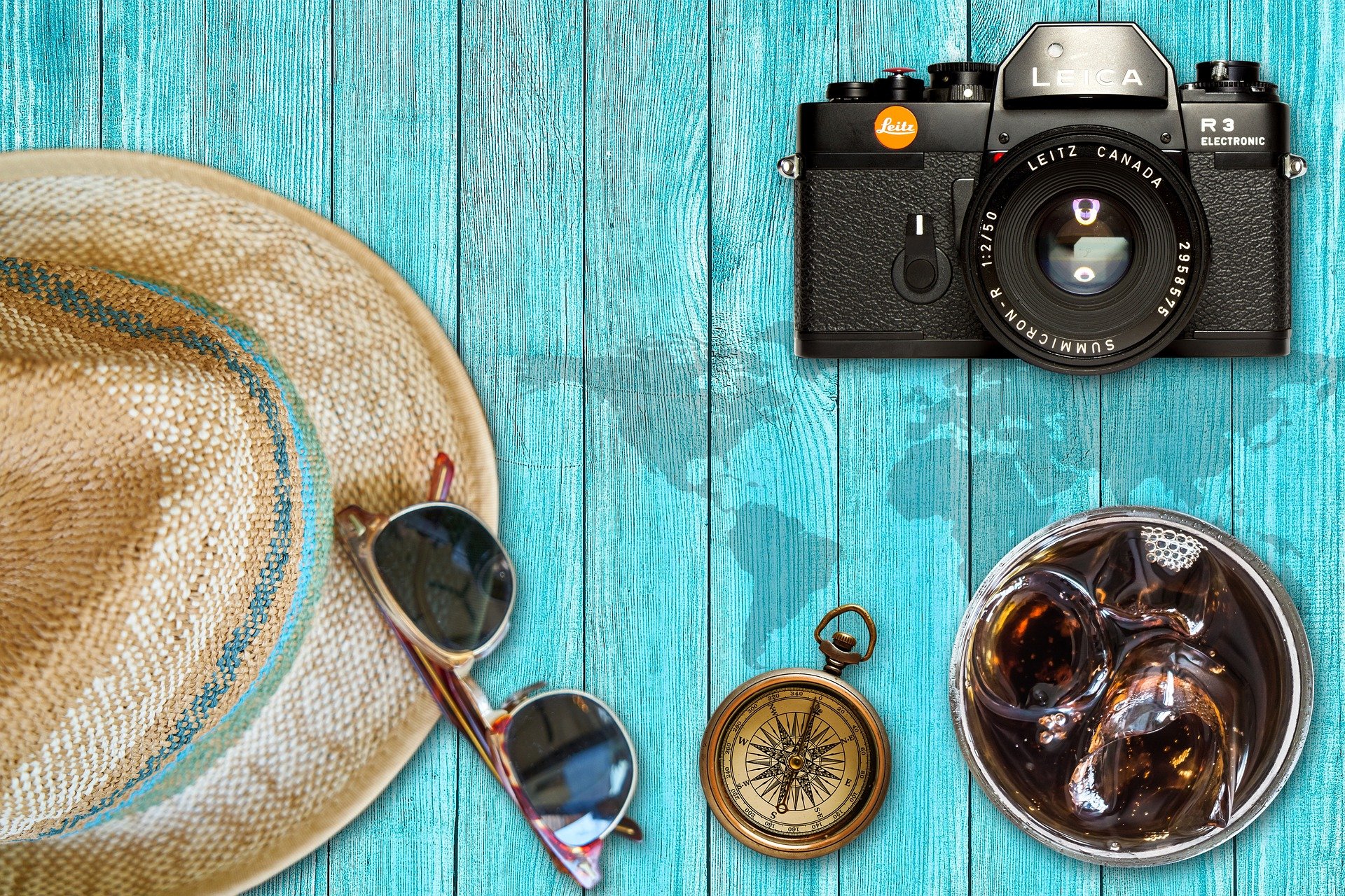 Foto: Auf Tisch liegen Kamera, Sonnenbrille, Strohhut, Kompass, Getränk mit Eiswürfeln