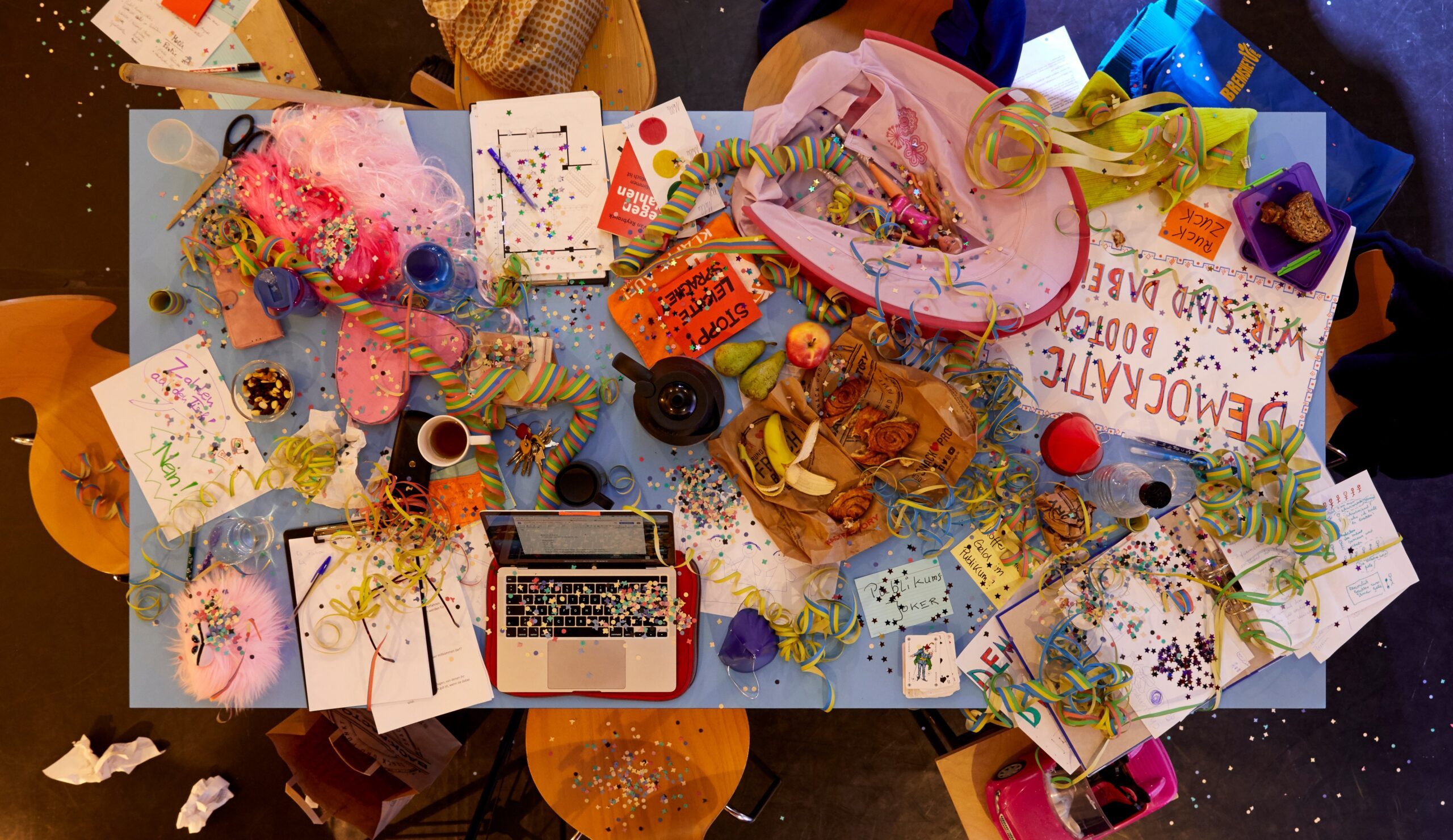 Tisch von oben: ein buntes Durcheinander aus Laptop, Luftschlangen, Obst, Kaffeebechern