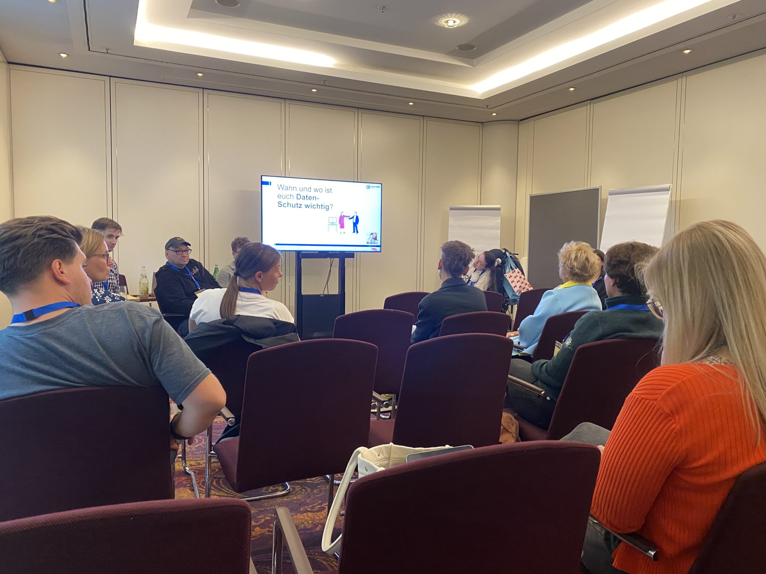 Ein Publikum von hinten zu sehen, Ein Bildschirm mit Präsentation vorne und vortragenden Menschen.