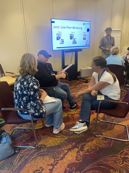 drei Menschen in einer Sitzgruppe, Bildschirm im Hintergrund