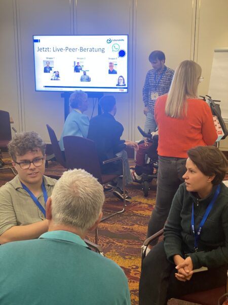 Im Vordergrund drei Menschen die zusammensitzen und sich unterhalten, im Hintergrund ein Bildschirm und weitere Menschen