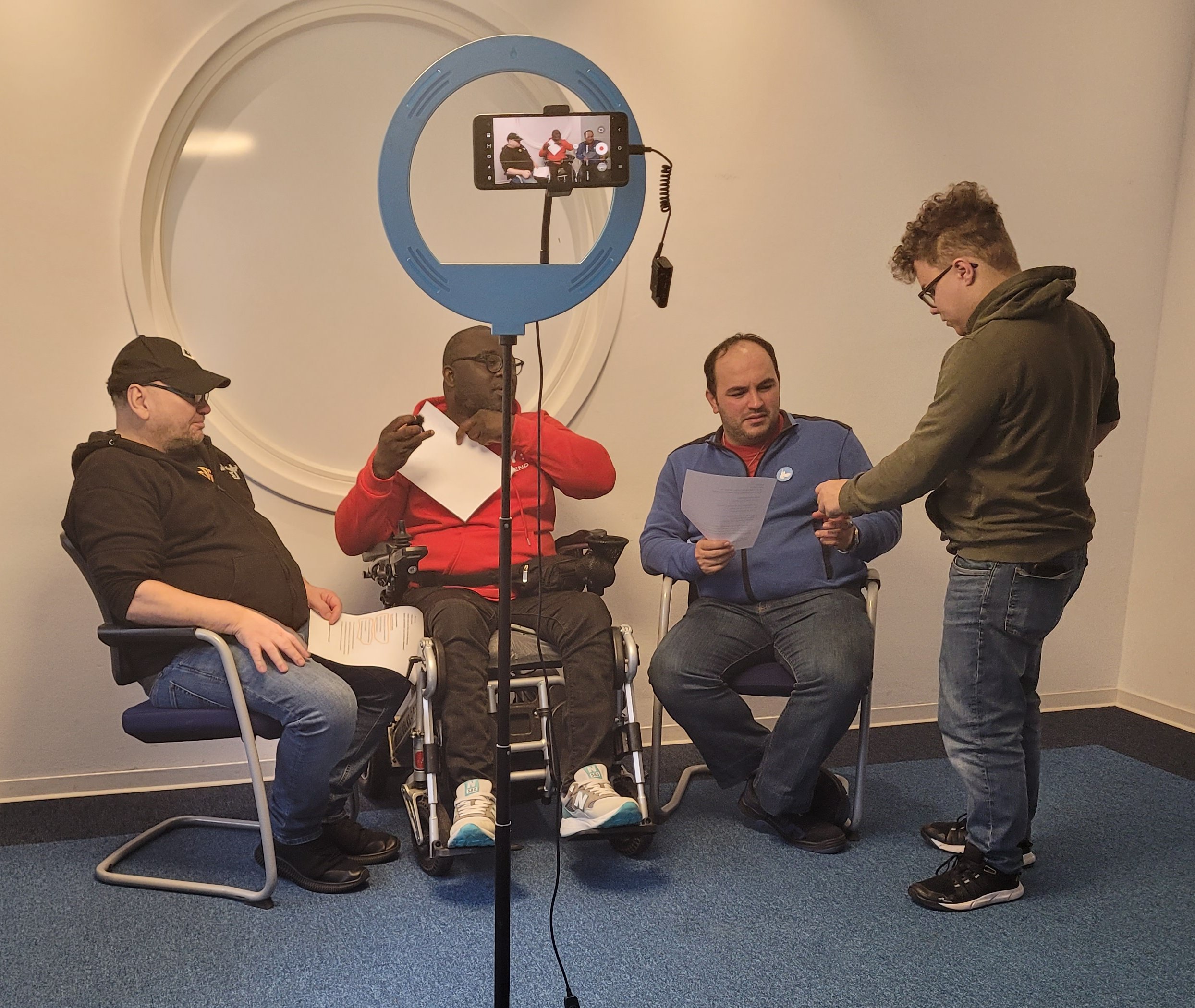 Drei junge Männer, davon einer im Rollstuhl, sitzen vor einem Ringlicht.Ein Mann steht daneben und erklärt etwas.