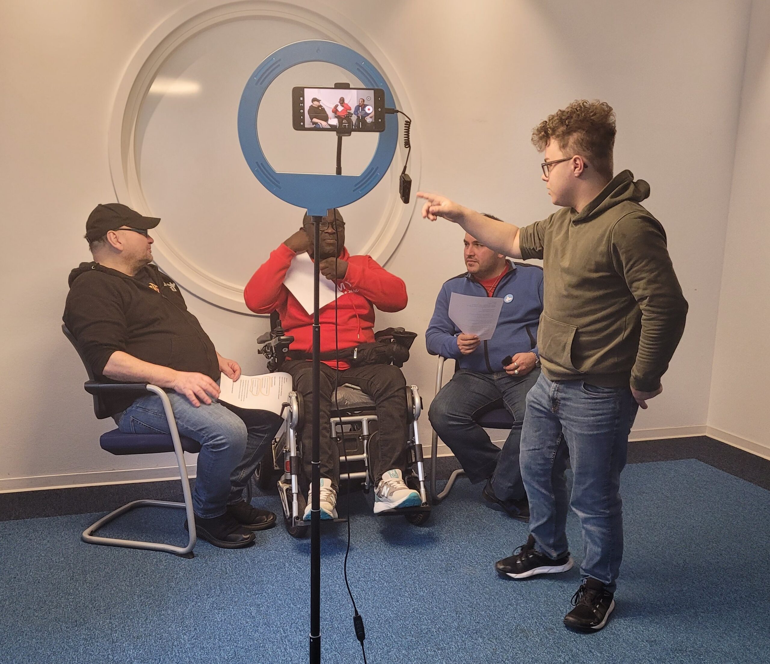 Drei junge Männer, davon einer im Rollstuhl, sitzen vor einem Ringlicht.Ein Mann steht daneben und erklärt etwas.