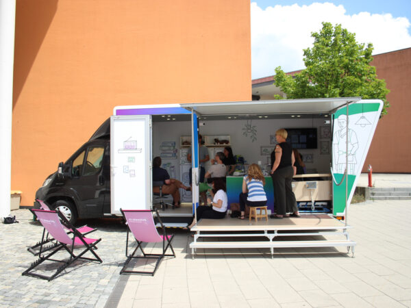 Ein seitlich geöffneter Truck steht auf einem sonnigen Hof. Im Truck schauen Menschen sich Technologien an.