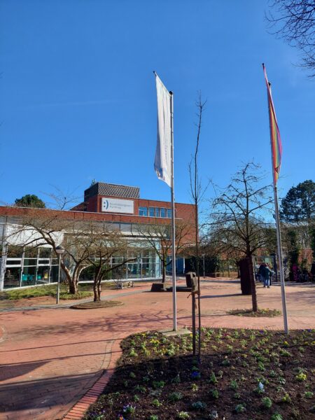 Ein Schulhof mit Bäumen und zwei Fahnen, das Gebäude im Hintergrund