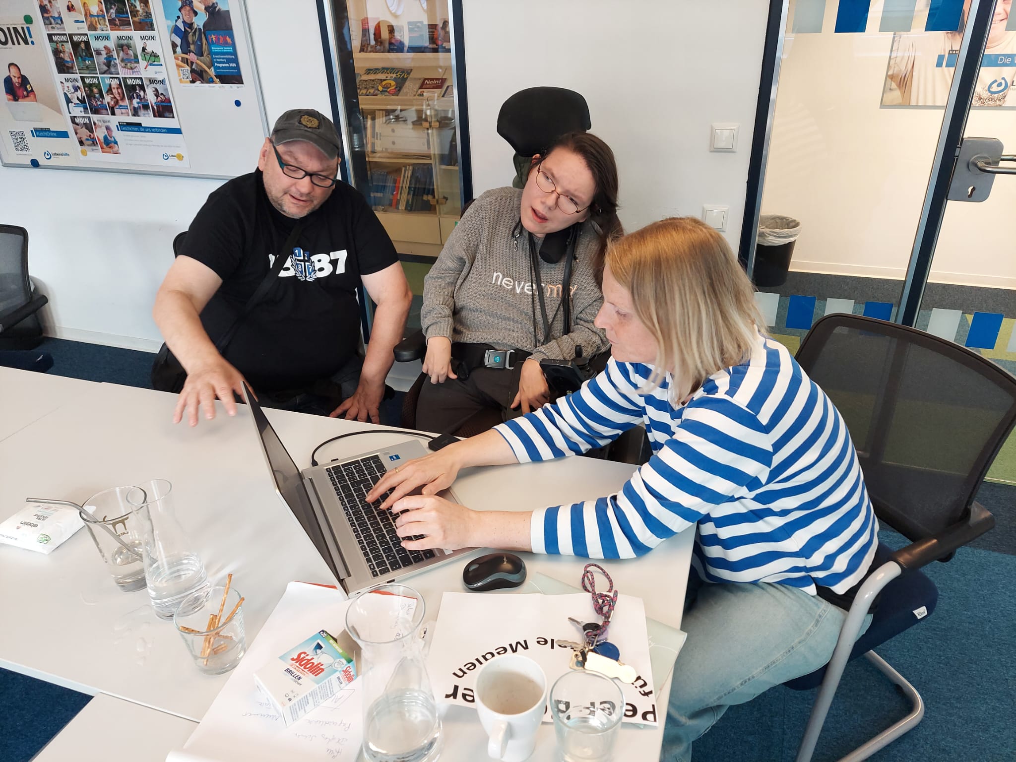 Frau tippt auf Laptop, im Hintergrund schauen zwei Menschen zu.